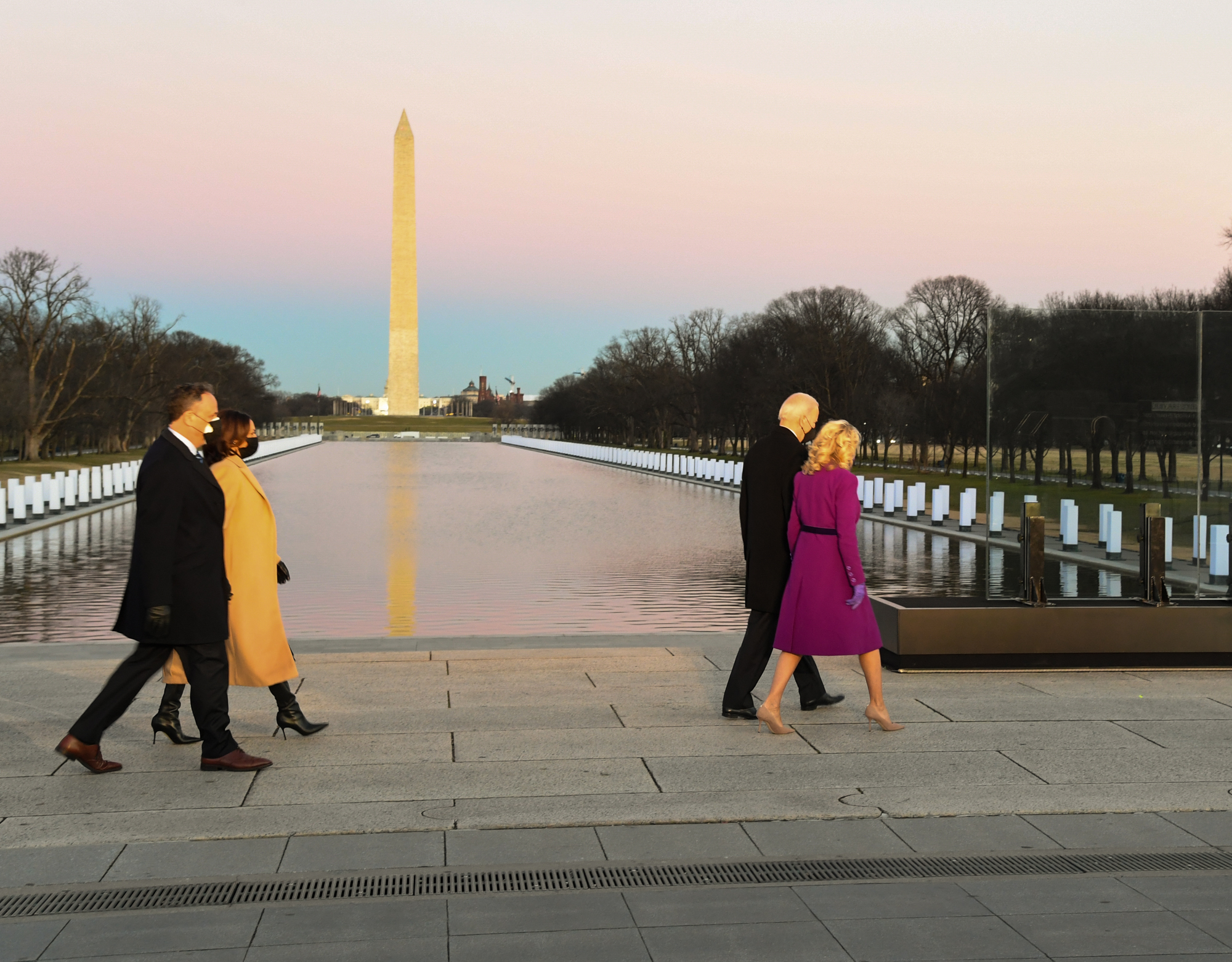 Joe and Jill Biden, Kamala Harris, and Doug Emhoff walk next to a reflecting pool lined with lanterns at dusk. The Washington Monument, US Capitol building, and spotlight are in the distance. 