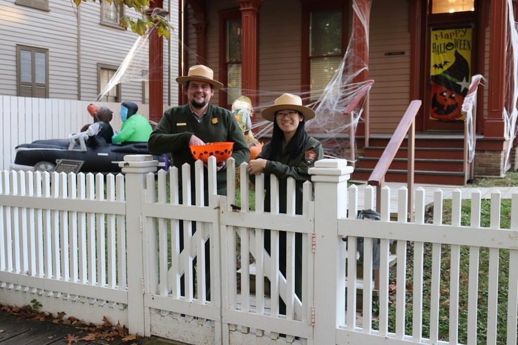 Two park rangers, in green uniforms and straw hats, stand in front of a white picket fence and wooden house holding an orange candy bowl.