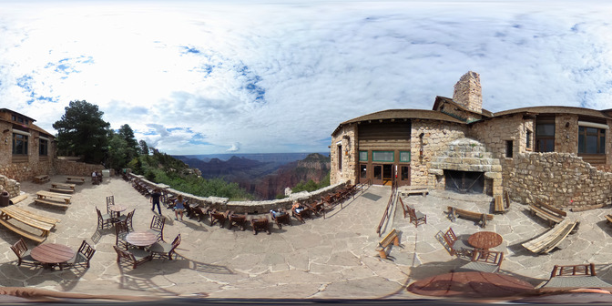 The flagstone patio of a rustic lodge has a long row of wooden deck chairs along a stone guard wall that overlooks the vast Grand Canyon landscape. The stone walls and fireplace of this rustic lodge wrap around the flagstone patio and several picnic tables are placed away from the overlook itself. 