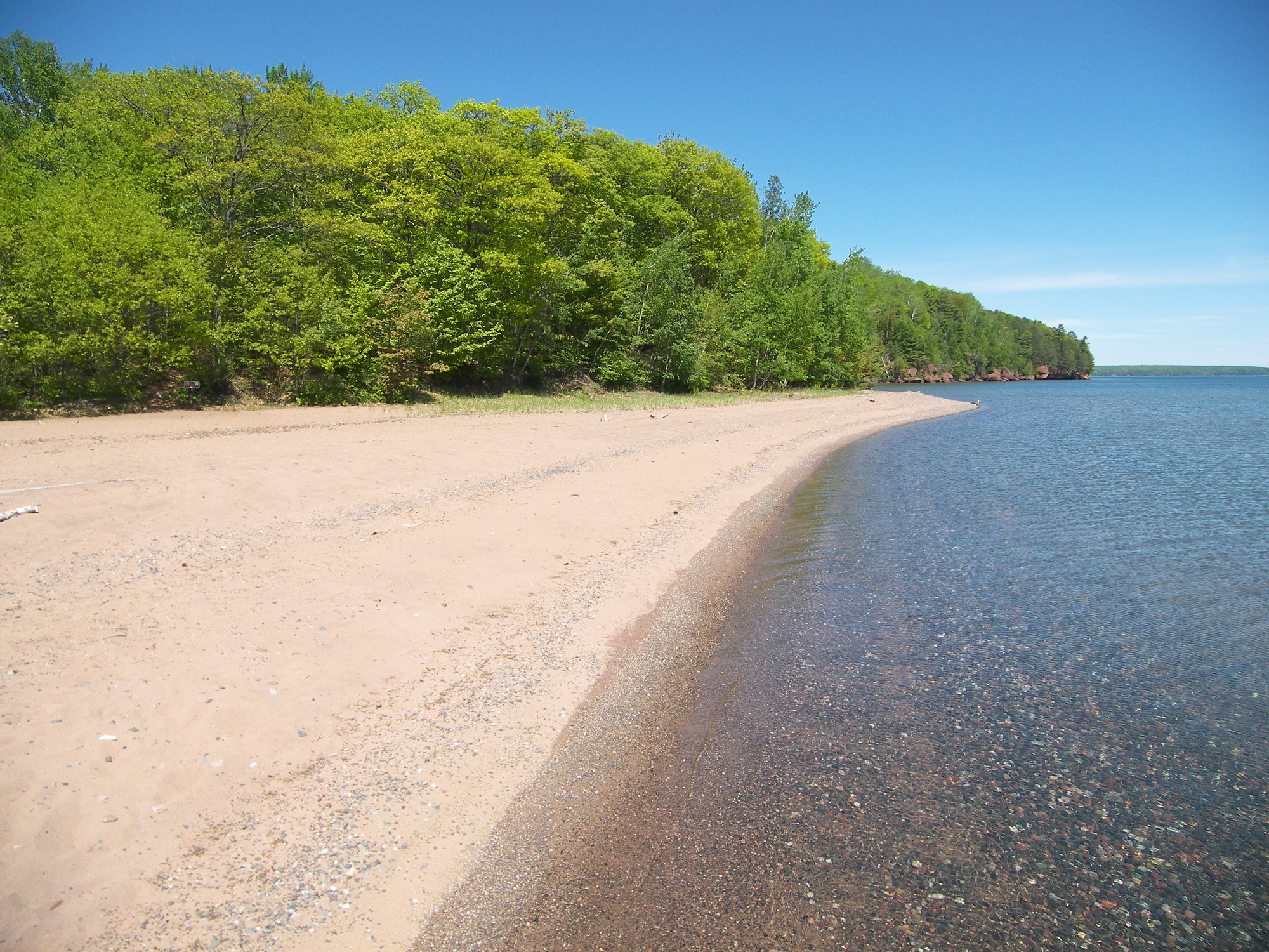 An empty sand beach surrounded by trees on one side a clear water with rocks on the other. 
