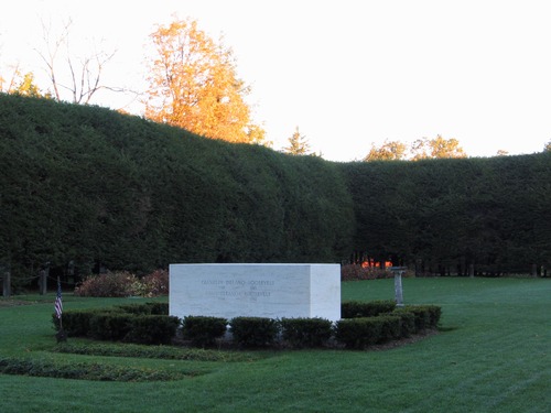 Garden surrounded by a tall hedgerow. White marble stone burial marker.