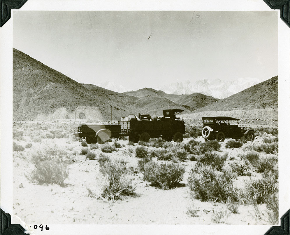 This is an historic black and white photograph from the Scotty's Castle Historic Photograph Collection, Death Valley National Park of passenger vehicle driving towards right edge. Followed by large truck pulling supply trailer. Truck full of supplies. Parked on desert floor. Barren hills beyond flats. Faint snow-covered mountains in back. Number in black ink in lower left corner.