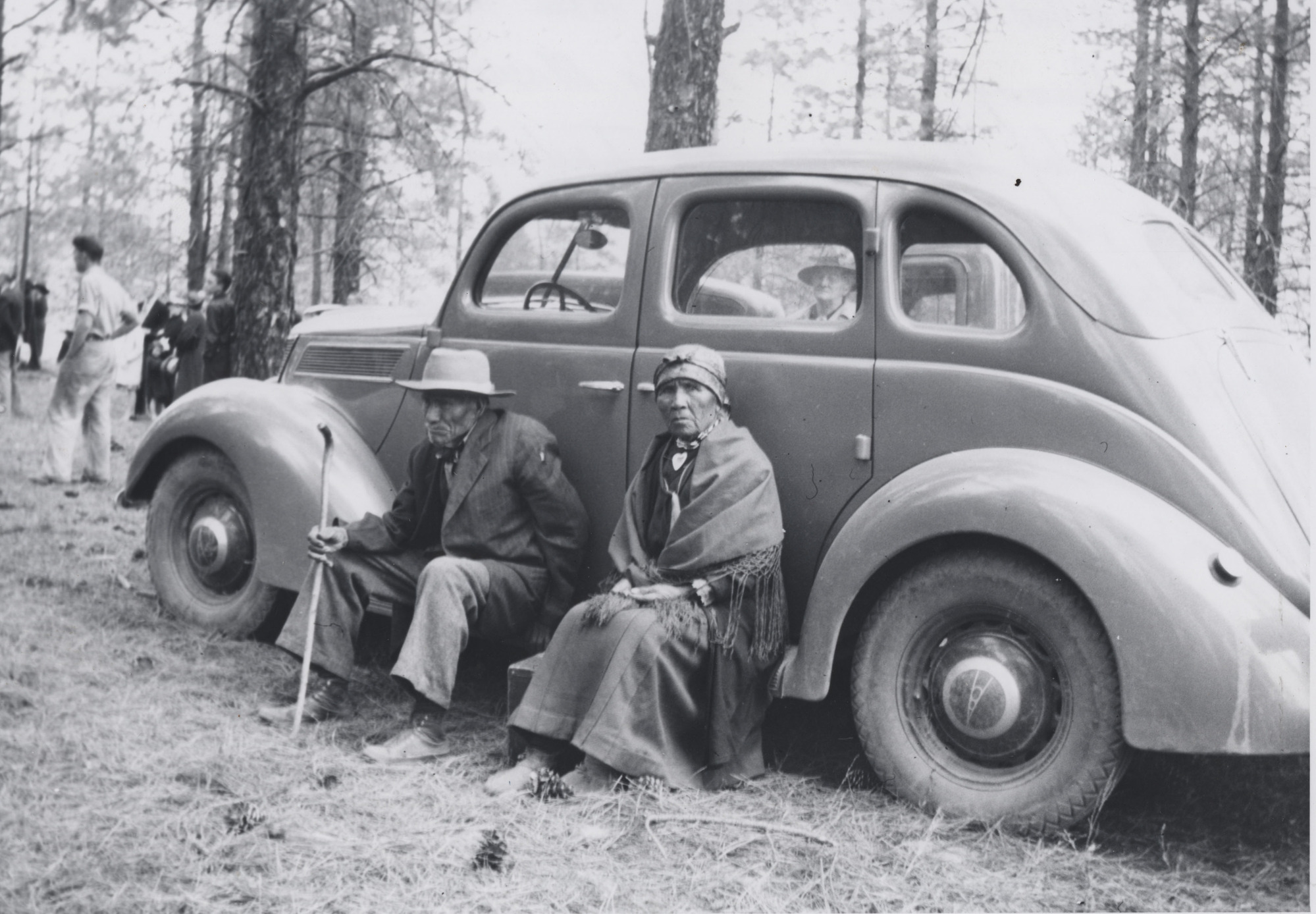 Black and white photograph of an elderly man and woman sitting on the step of a car
