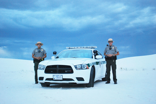 Two law enforcement rangers stand next to a patrol car out in the park.