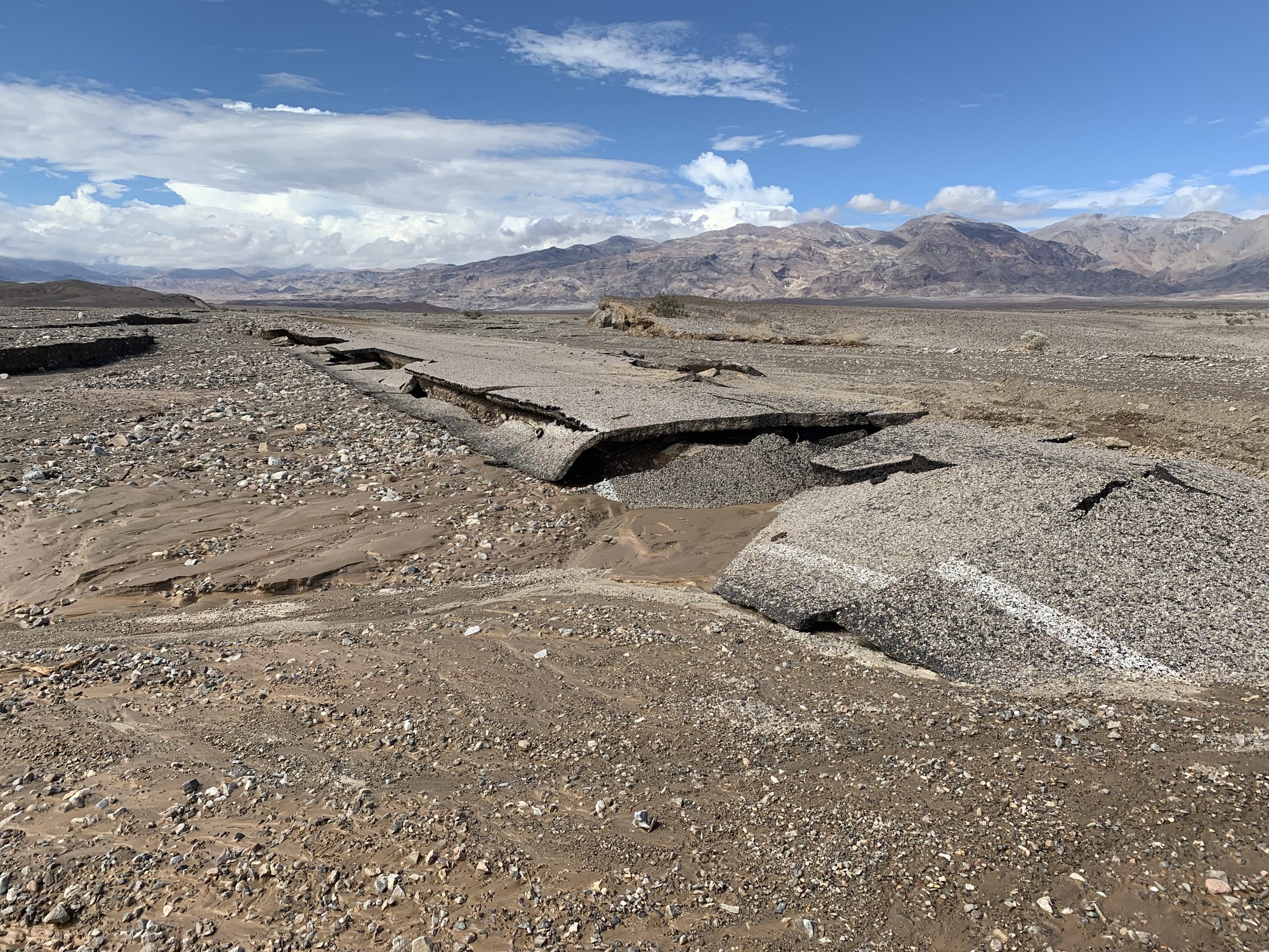 Mud and rocks left behind from flash flood in the foreground, asphalt broken in places behind it in front of a mountain landscape.