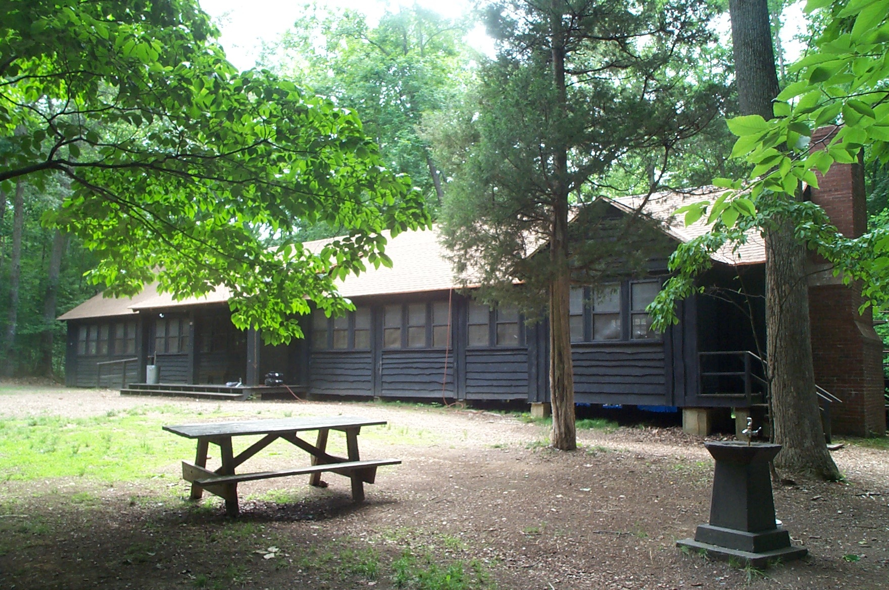 Large wooden cabin building with many windows and a picnic table and water fountain out front at the edge of a green forest