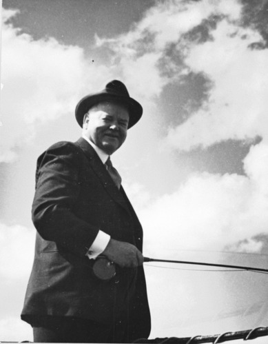 A black and white photo depicts a smiling middle aged man in a hat outdoors.