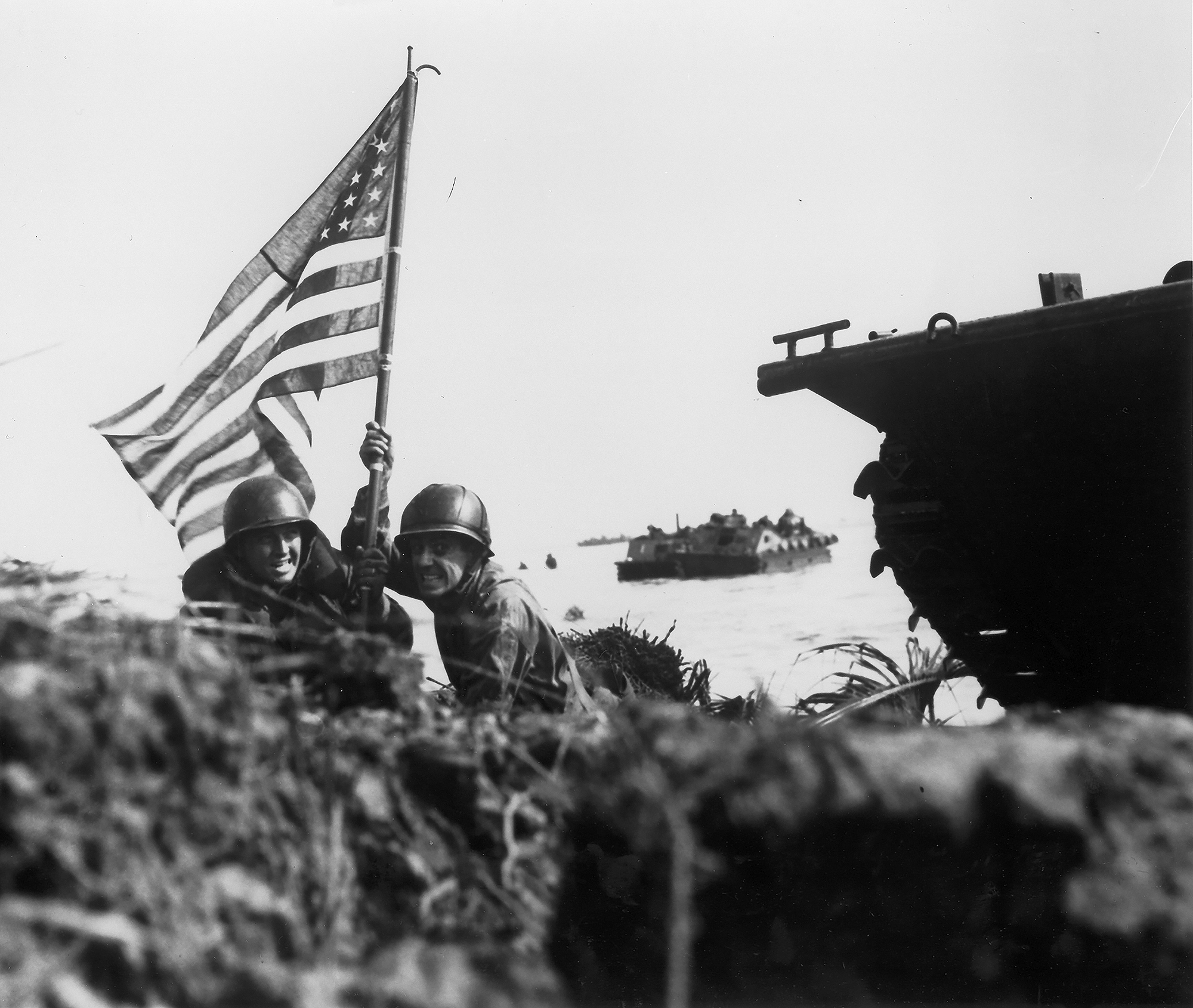Two soldiers hold an American flag hoisted on a boat hook during a battle.