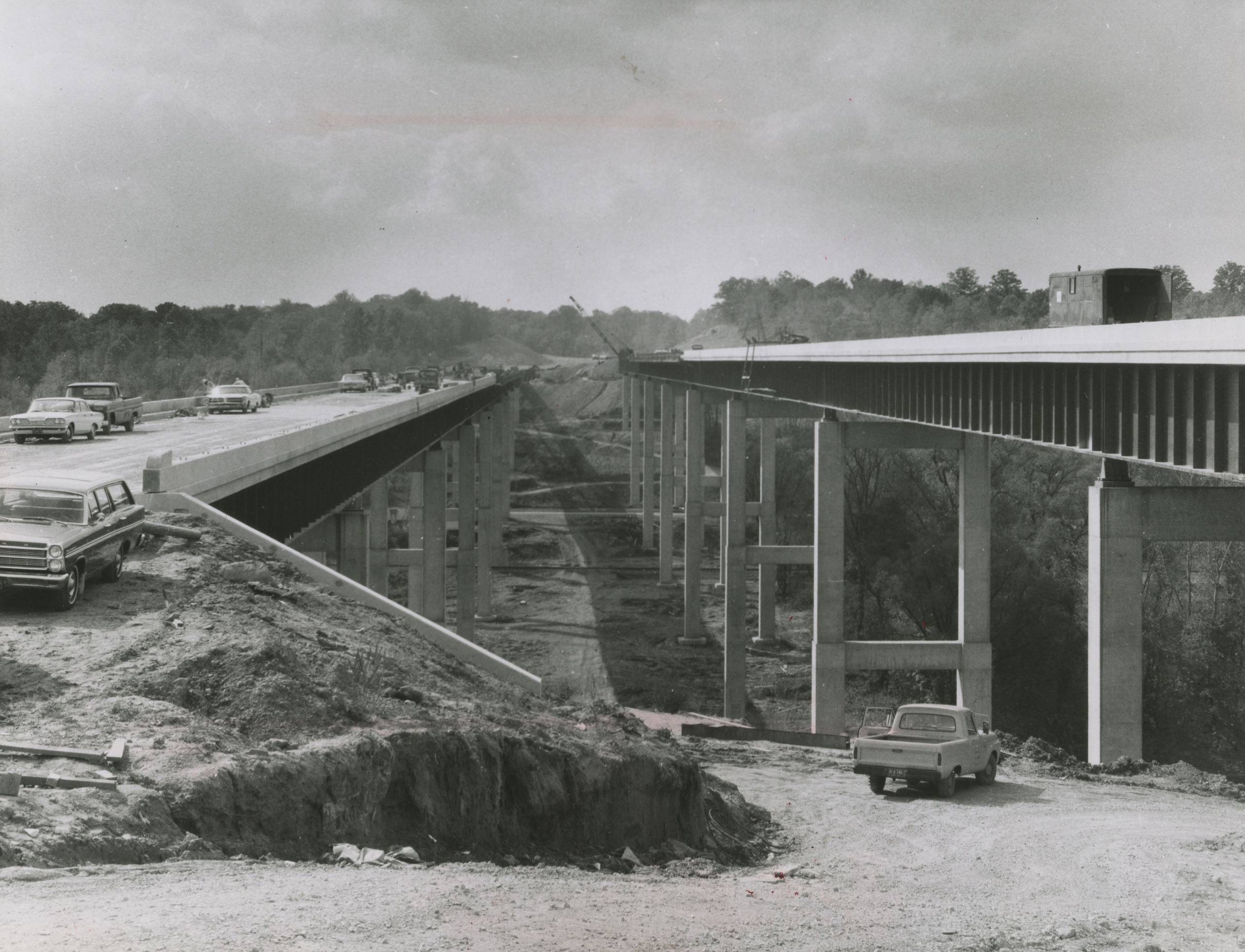 Work vehicles and equipment are parked on two concrete and steel highway bridges that cross high over a valley.