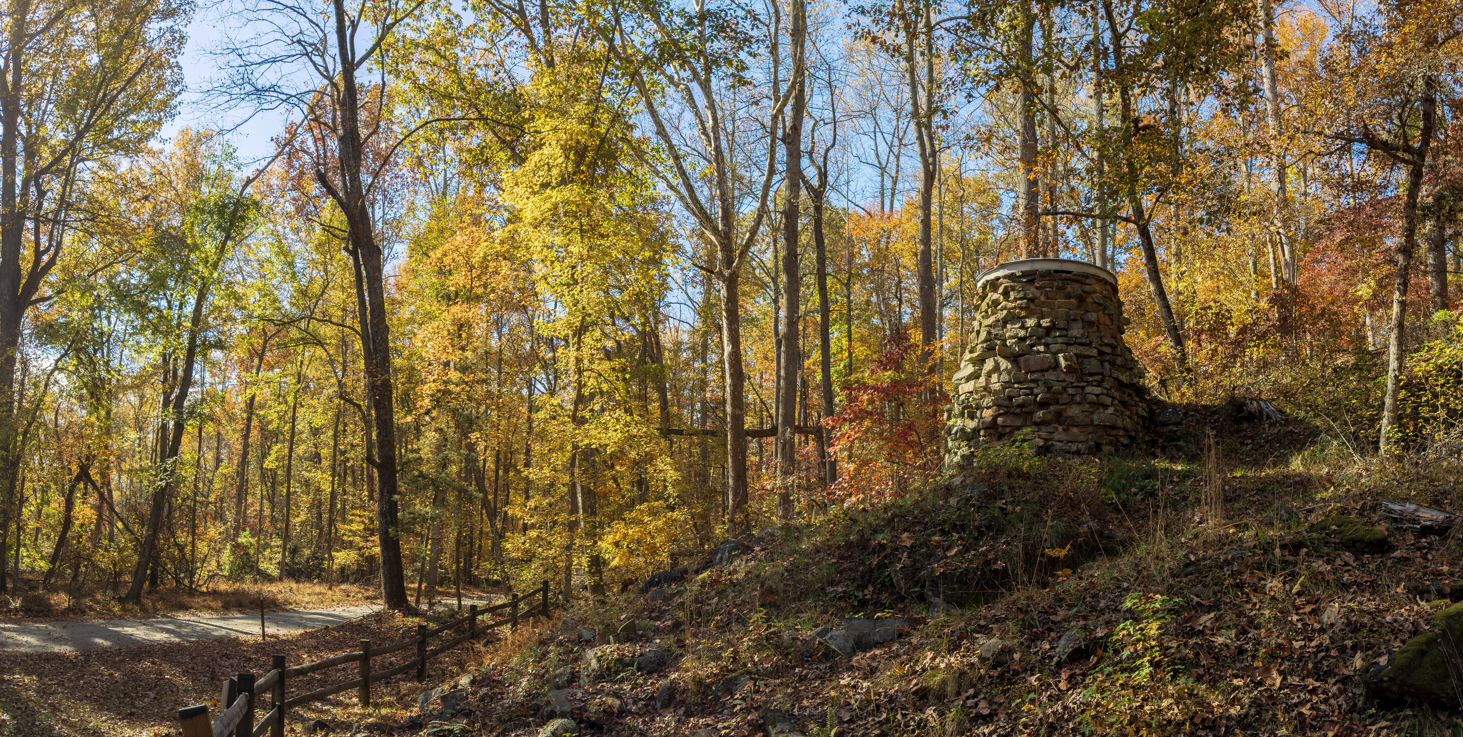A stone cylindrical ruin of a furnace stack on a rocky knoll surrounded by trees turning fall colors. 