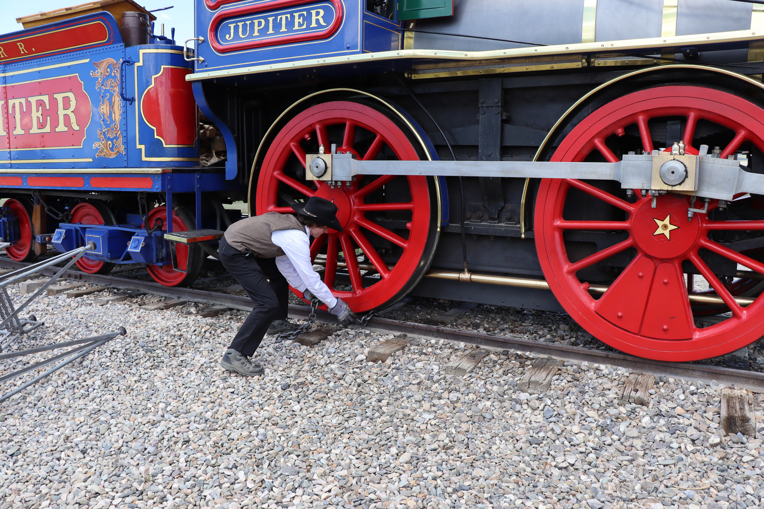 Locomotive fireman places a safety chain around the wheel of Jupiter a blue and red 1860's Victorian steam locomotive. 
