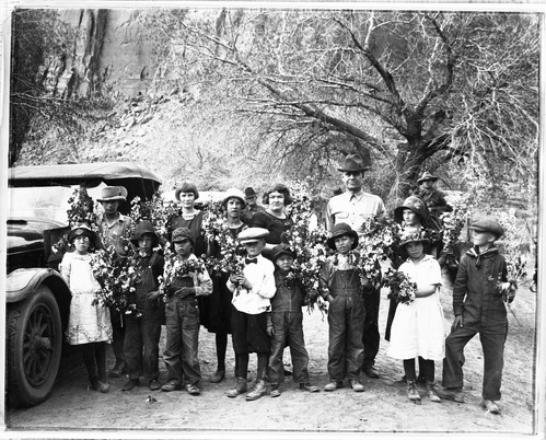 Man in hat, women, and many children with garlands of flowers stand by an old car, below cliffs. 