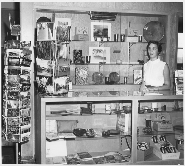 Black and white. A woman standing behind a shop counter. With shelves of items for sale and postcard racks off to the left