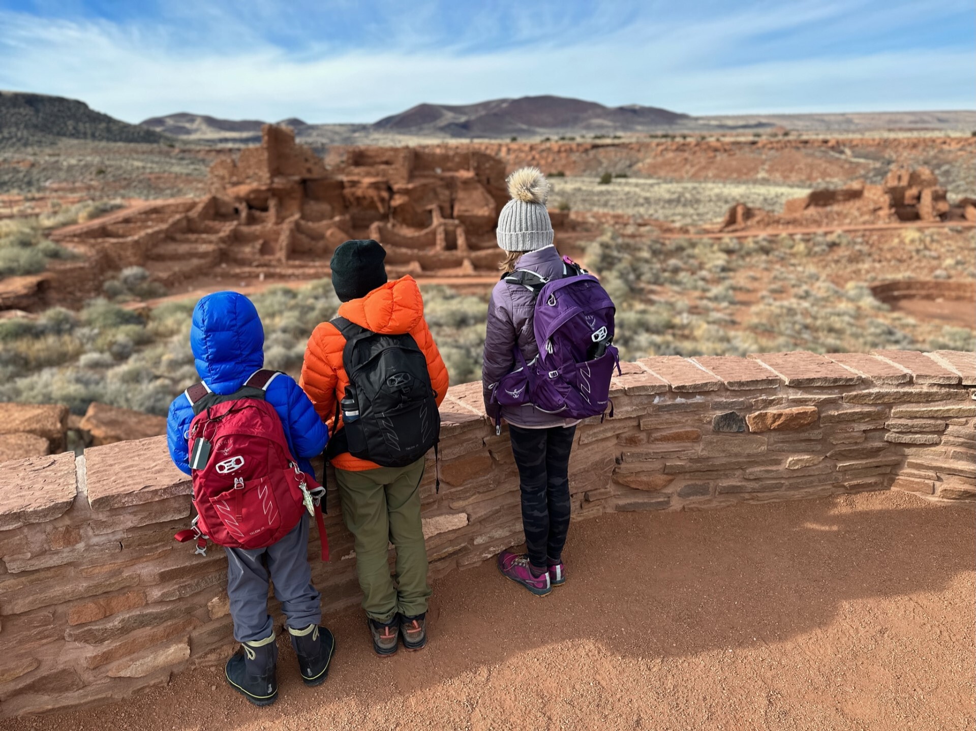 Three Jr. Rangers wearing cold weather clothing stand at the Wupatki Pueblo overlook. 