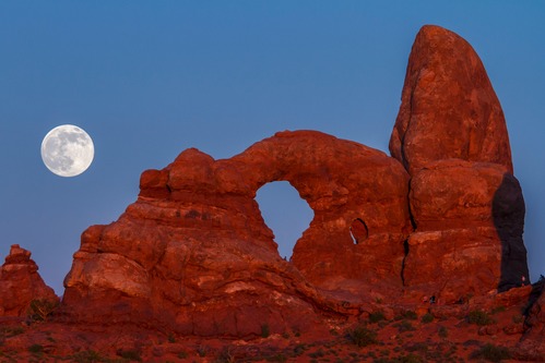 a full moon rises above a red-rock arch