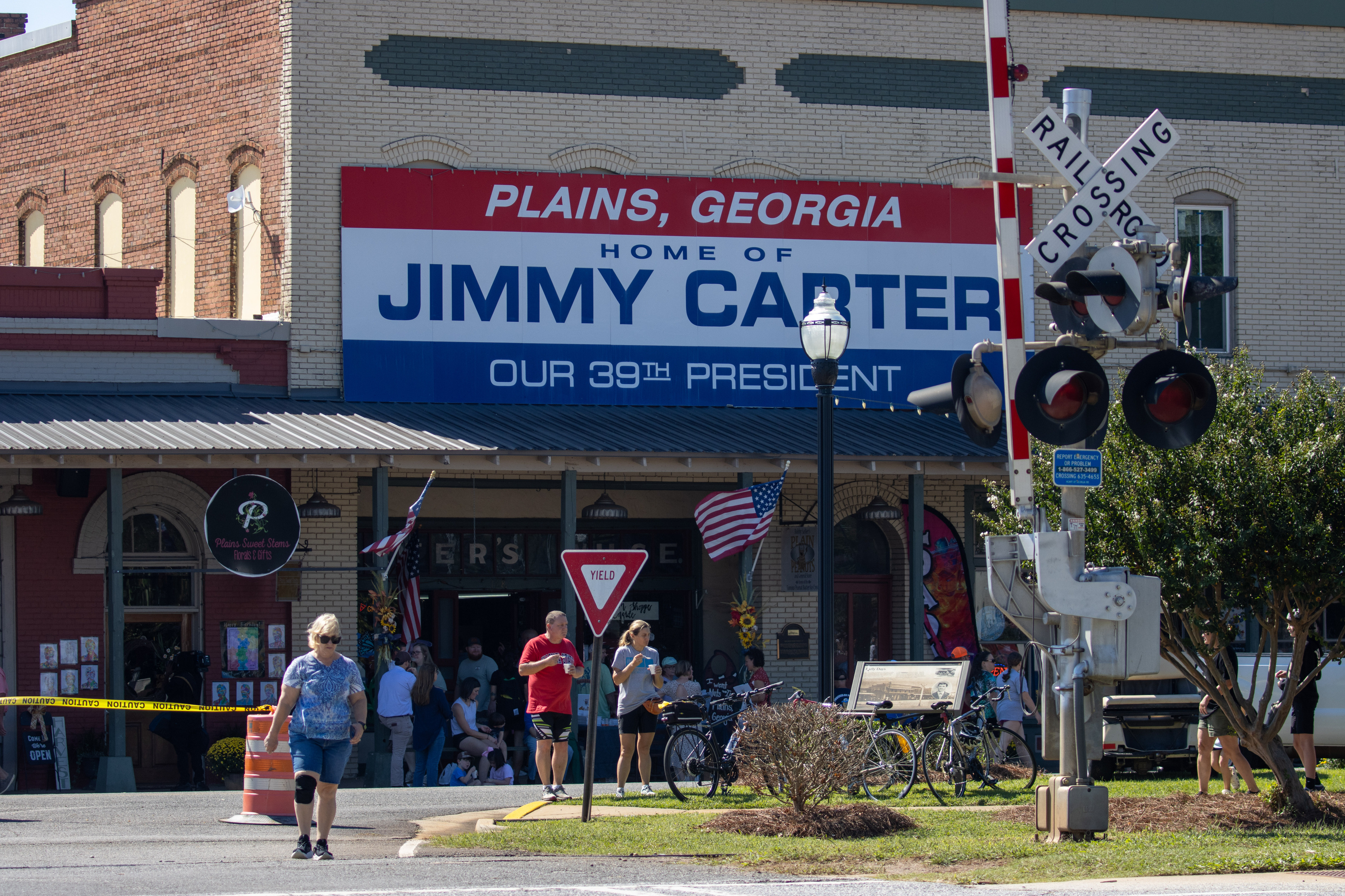 Downtown Plains, G.A. with crowd of people featuring a building that has a large sign, "Plains, Georgia. Home of Jimmy Carter our 39th President" and a railroad crossing sign. 