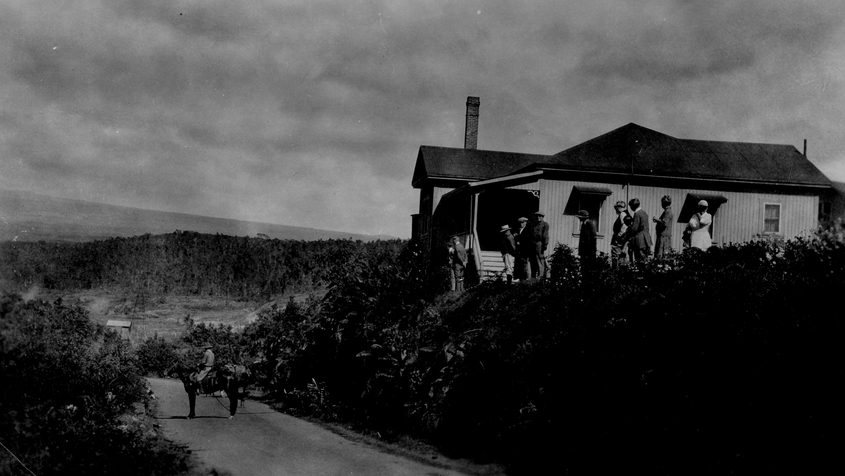 Black and white photograph of the side view of a house on a small hill above a road. The road is present towards the left side of the image and the house sits on the right side. In the road, towards the bottom, a man on horseback dressed in National Park Service attire faces away from the camera, and it is assumed they are conversing with another person in the road, as evidenced by feet below the horse and a dog next to them. Slightly above the road, on the side of the house, approximately ten men and women stand, and either looking towards the camera or towards the man on horseback. Lush vegetation is present around the house and to the left of the road. In the distance, Mauna Loa can be seen. The sky is fully covered in clouds.