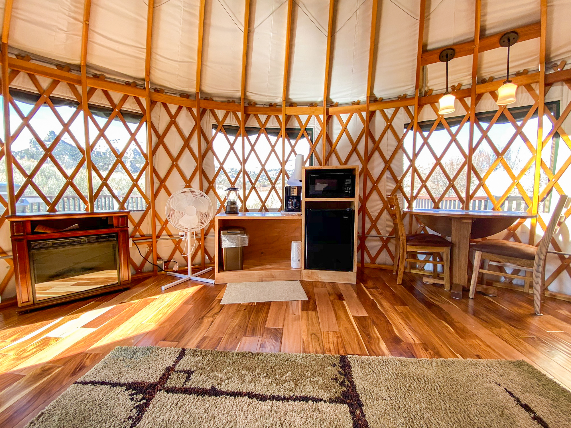 Mini fridge, microwave, coffee station, table with two chairs, fan, and fireplace style heater. Round yurt structure. It has a clear dome on the ceiling, exposed wood beam ceiling and exposed wood lattice wall supports. 