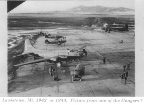 Black and white photo of tarmac with a large plane in the foreground, a few smaller planes and mountains in the background. People and trucks are visible on the tarmac. 