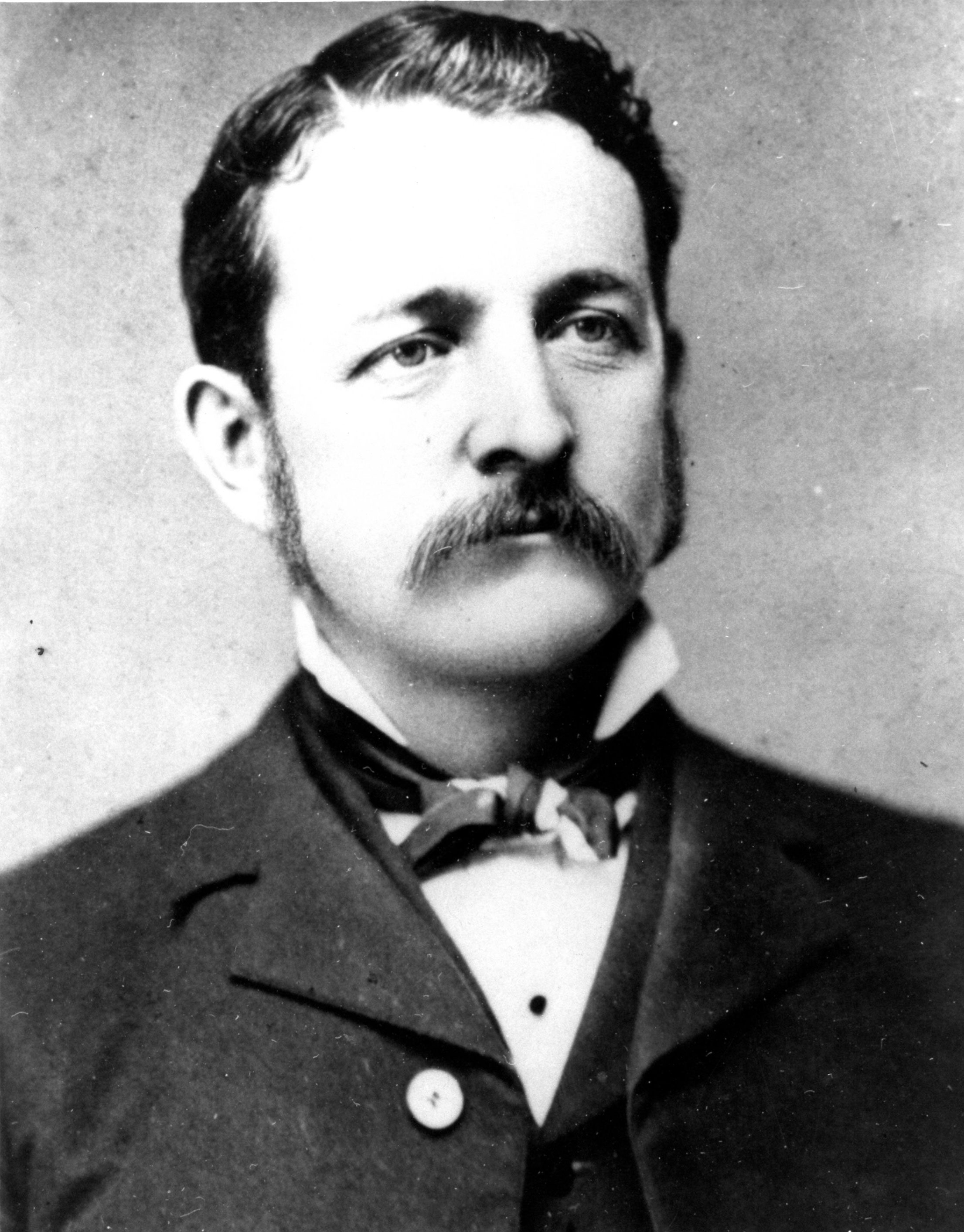 An 1883 portrait photo depicts a man with a mustache and sideburns in a suit with a bow tie.