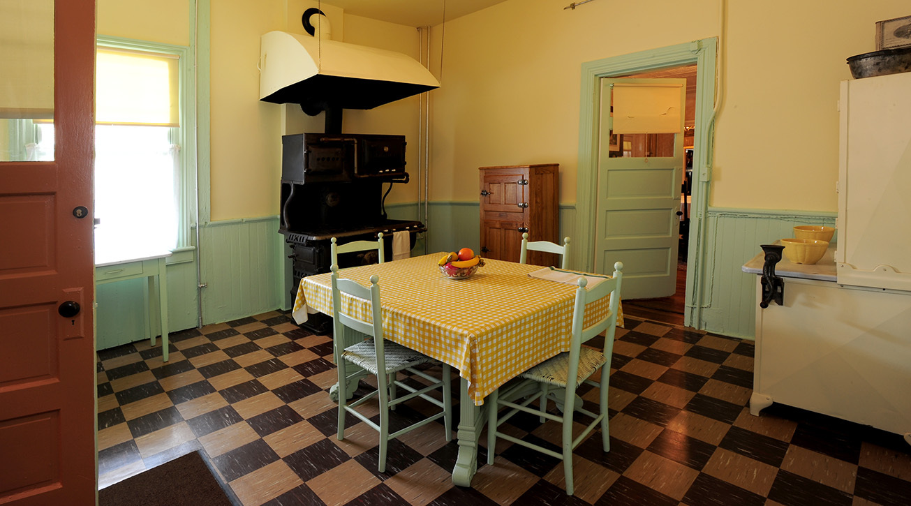 A Kitchen with a square table in the middle, stove and icebox and a Hoosier cabinet lining the room.