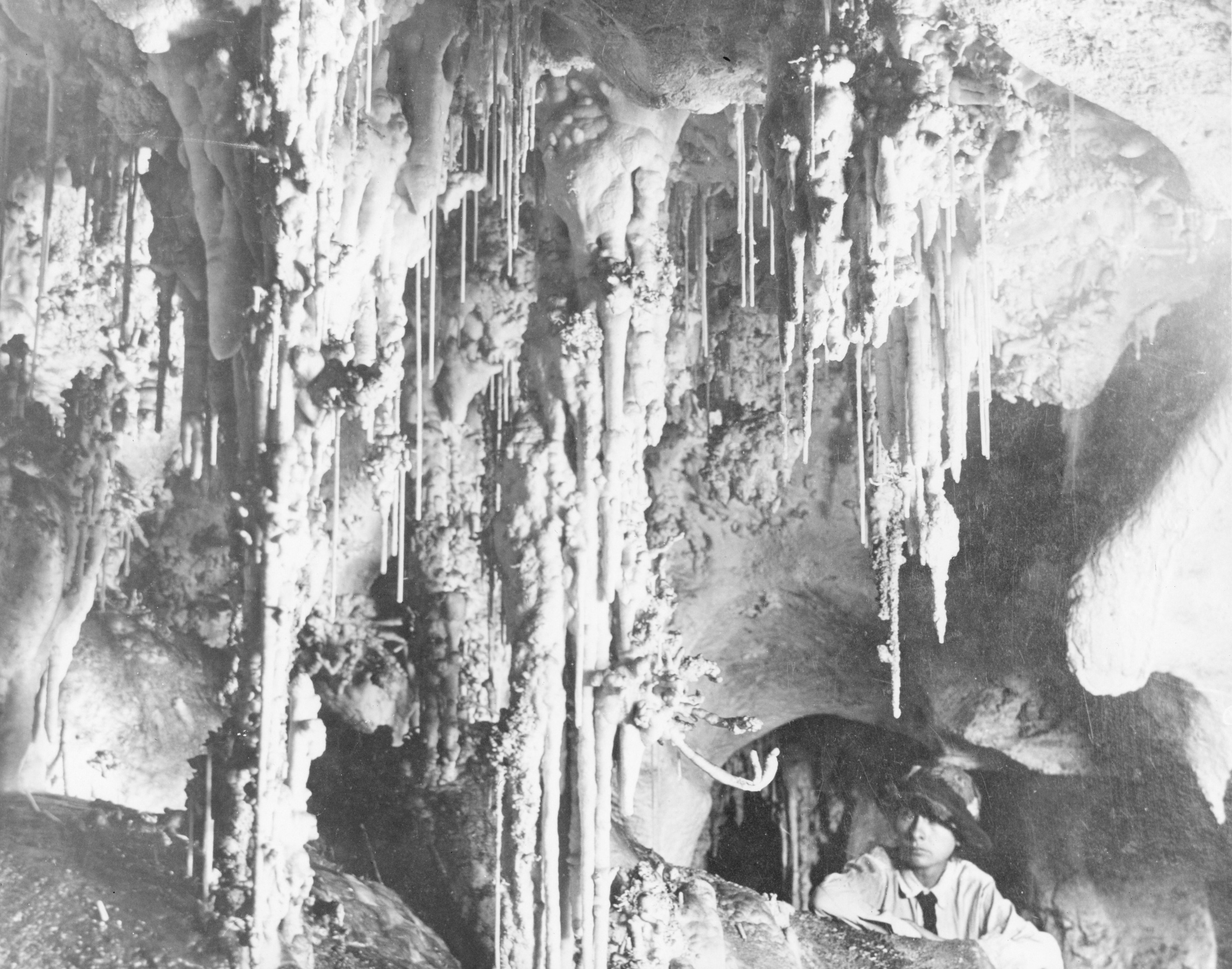 A black and white photograph of a person looking out at the many small and large speleothems from the ceiling. 