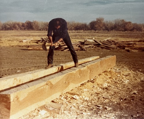 A man stands astride two long wooden beams and uses a hand tool to shape the wood. 
