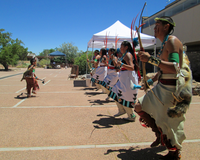 Row of male and female dancers facing a boy dancer in a plaza with canopies, trees and blue sky in the background.
