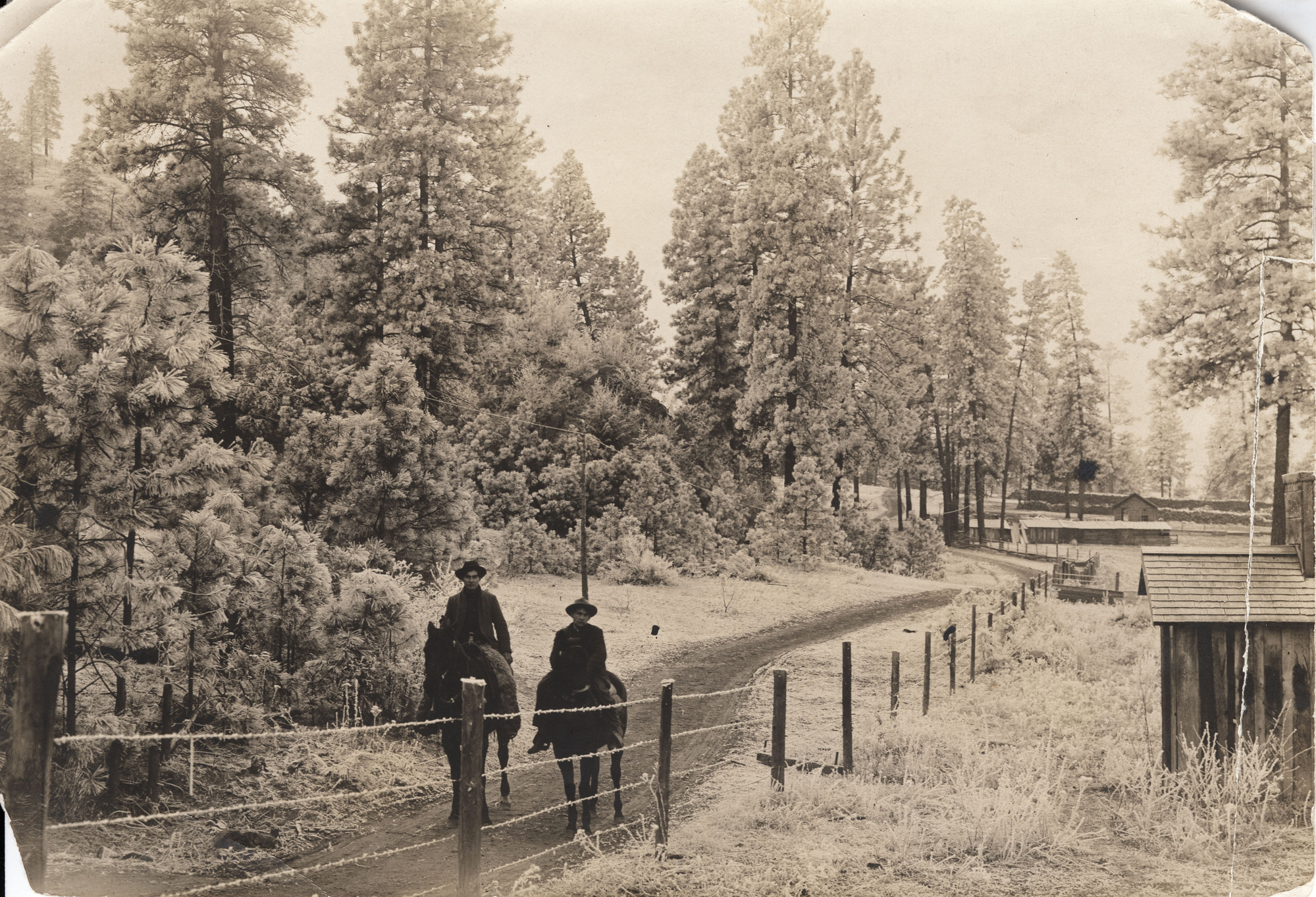 Black and white photograph of a frosty scene with two people on horseback among buildings and trees