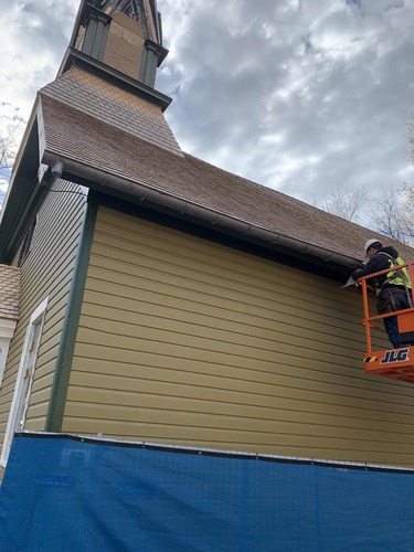 A construction worker stands on a lift at the side of the church and adds a section of metal gutter along the bottom edge of the roof.