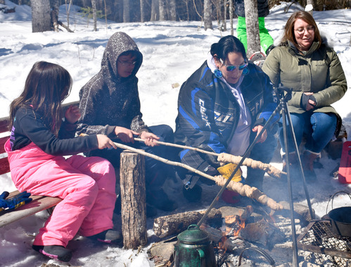 Two young people and two adults are seated near a campfire in a snowy landscape. The young people hold sticks with dough wrapped around the ends over a fire.