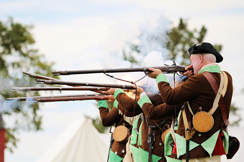 Men dressed in brown jackets with green insides with black hats fire muskets as white smoke and sparks billow out of them. 