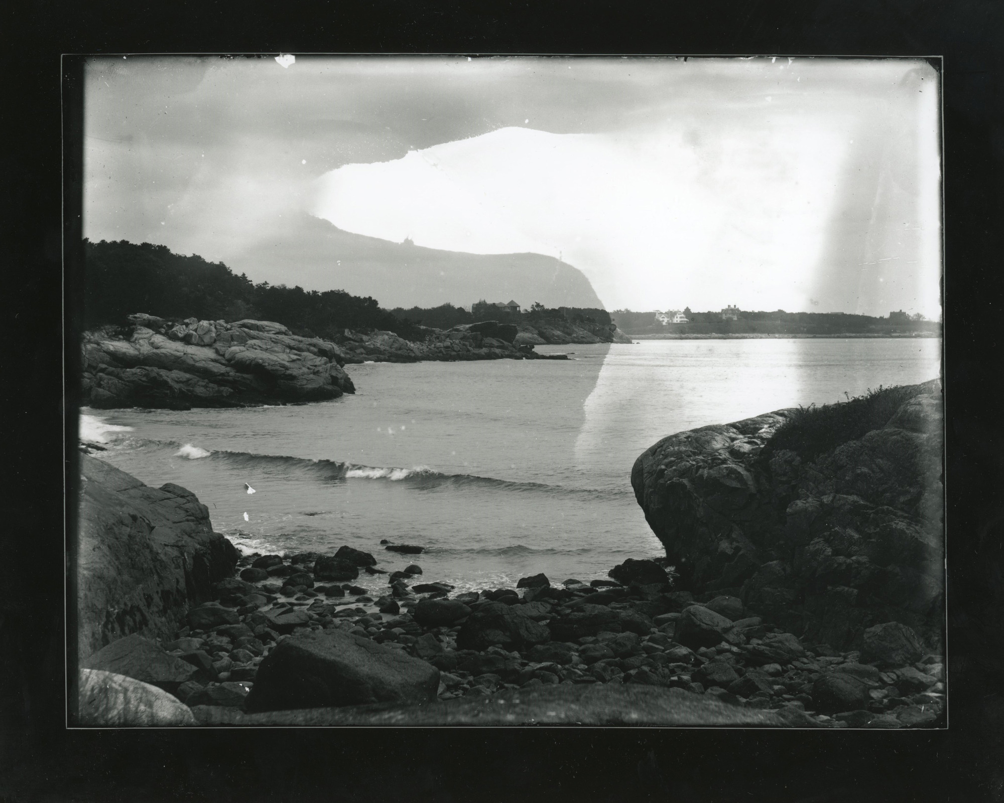 A rocky beach meets water. Trees and houses on land in background.