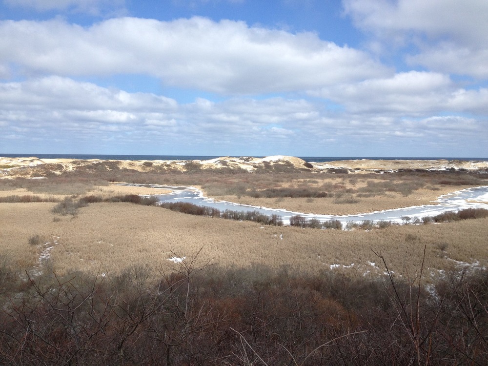 Beige sand dunes appear as undulating hills stretching to the water, while white clouds drift overheard.