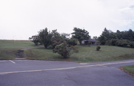 A building, partially hidden by trees, stands on the side of a grassy slope above a parking lot.