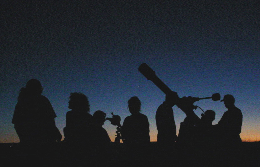 Seven people and a long telescope stand as a black silhouette from dusk's last light.