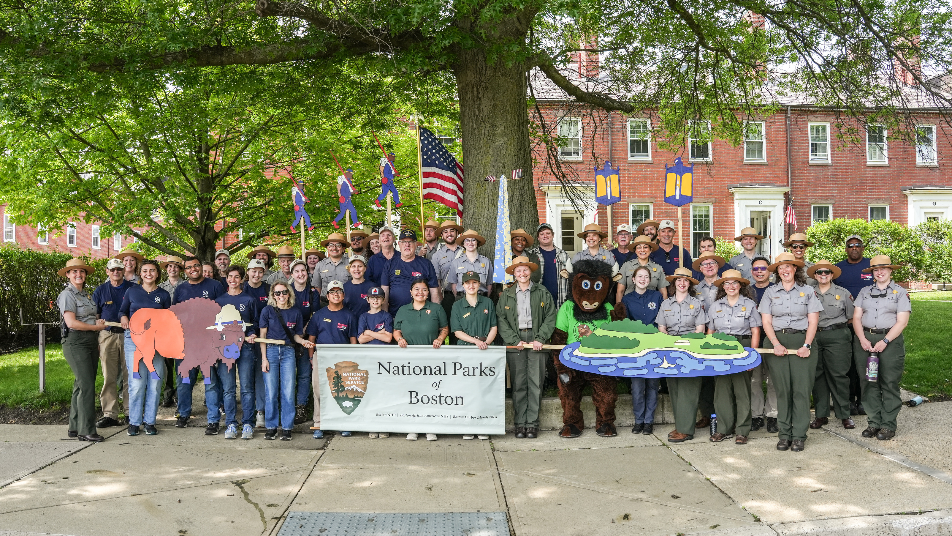 group of NPS staff, volunteers, SCA Historic Preservation Corps members, interns, and community members lined up with a National Parks of Boston sign and decorative wooden painted signs, "floaties," featuring different aspects of the park.