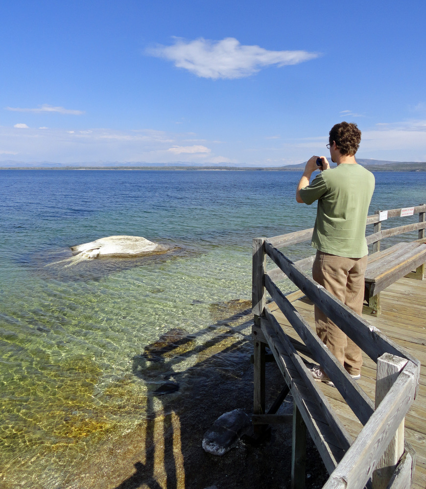 Man standing on boardwalk pointing camera at geyser cone sticking out of water in large lake