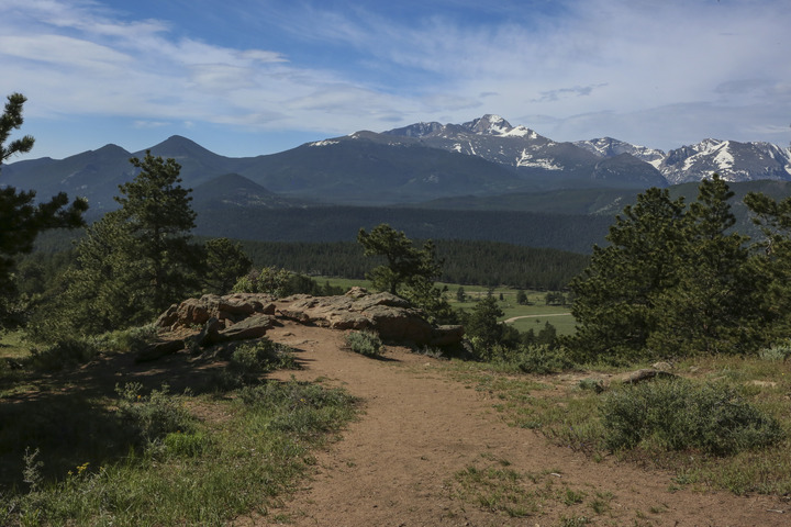 A dirt path leads to a rock with an expansive view over meadows, forests, and mountains.