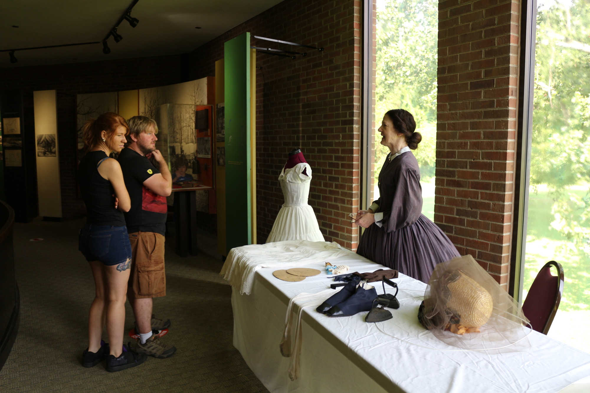 A man and woman look at a display of historic women’s undergarments while listening to a woman wearing a pale purple dress with a hoopskirt.