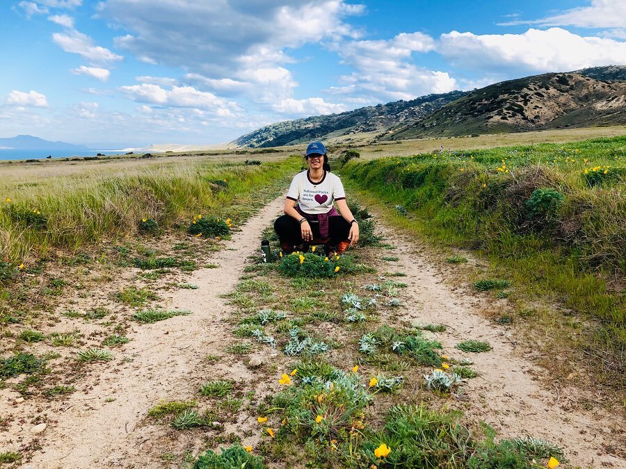 Visitor sitting in the middle of a trail with a giant blue sky and green mountains in the background