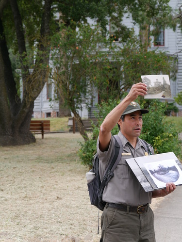 Keith Park in NPS uniform is holding up a photo of the John Muir Home, standing in the front driveway of the house. 