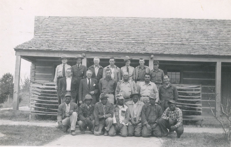 Seven African American men, and thirteen white men are in a group in front of a log cabin. Three of the white men are wearing ranger uniforms..