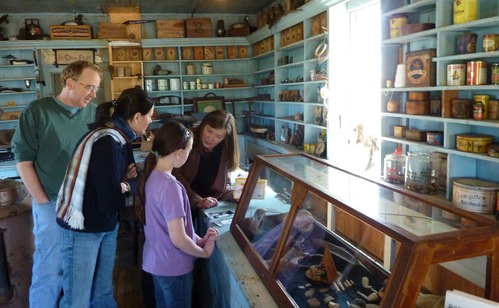 A family looks at goods for sale in a store with a long glass case in front and well-stocked shelves on the walls.