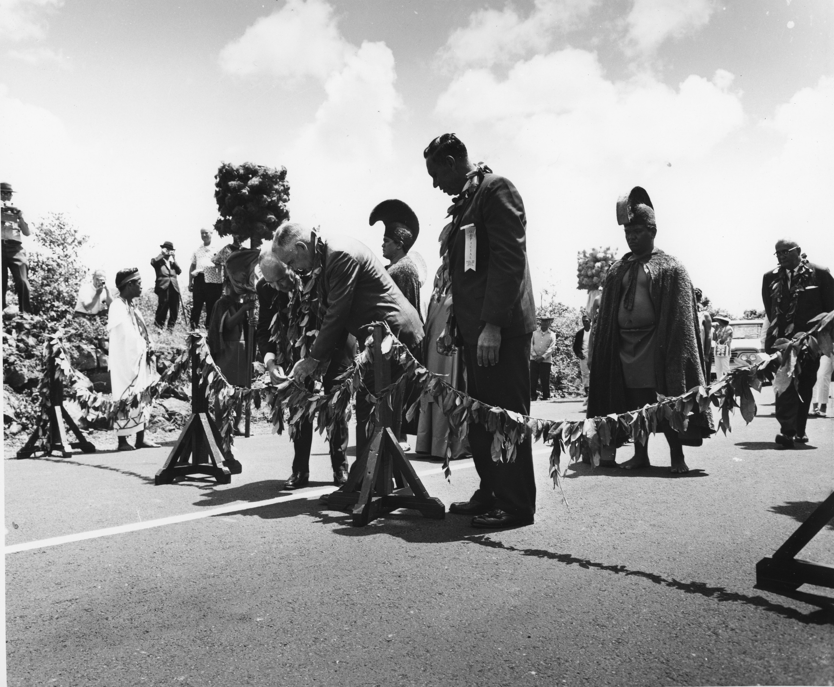 A black and white image of a dedication ceremony for the Kalapana section of Chain of Craters Road on June 9, 1965. A group of six people wearing suits and traditional Hawaiian clothing are standing on the newly paved Chain of Craters Road. Three men wearing suits have Ti leaf leis around their necks and are standing in the center of the image. One of the men wearing a suit has a “official” ribbon attached to the front of his jacket. To the left of him are the other two men wearing suits. They are both looking down at the road fixing the garland that runs across the road in front of them. In the background there is a group of spectators watching the dedication ceremony from the side of the road. Some of the spectators are holding up cameras to their faces.
