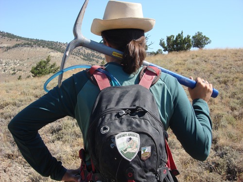the back of a woman wearing a straw hat and backpack with a pickax slung across her shoulders. 