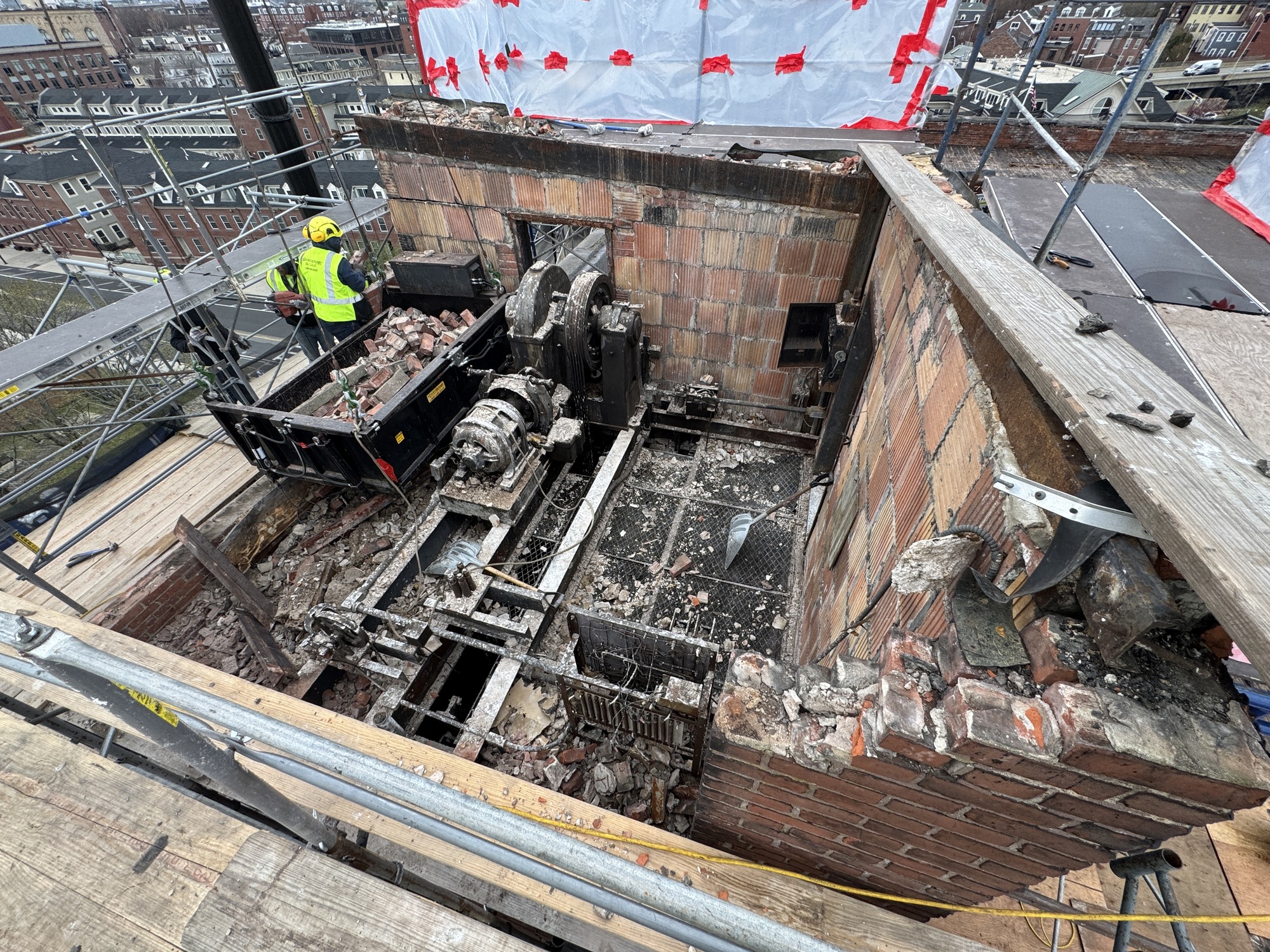 Debris of brick and other materials being removed from partly standing bulkhead on the top of a roof of an industrial building. 