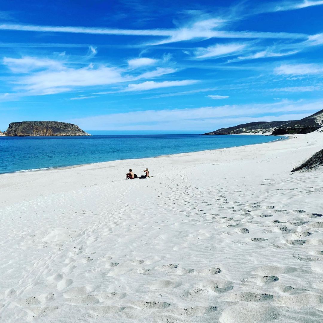 2 visitors sit off in the distance on a white sandy shoreline with beautiful blue ocean and sky in the background