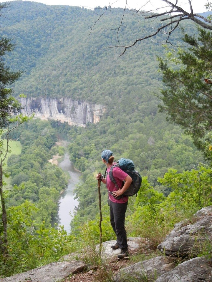 A hiker in a pink shirt with a blue bandana and backpack stands at an overlook of the Buffalo River above Steel Creek Campground.