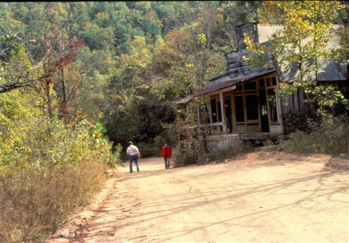 color photo of two people on dirt road near old wooden building.
